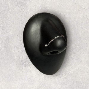 May include: A silver nose chain with a small, clear gemstone at the nostril end. The chain connects to a small ring that sits on the nose. The jewelry is displayed on a black face form against a gray background.