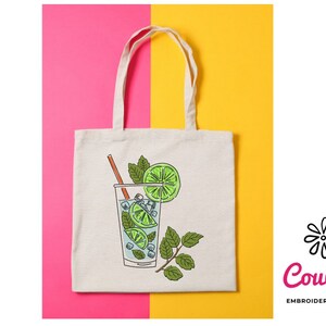 May include: A white tote bag with a drawing of a glass of mojito with lime wedges, mint leaves, and a straw. The bag has a pink and yellow background.