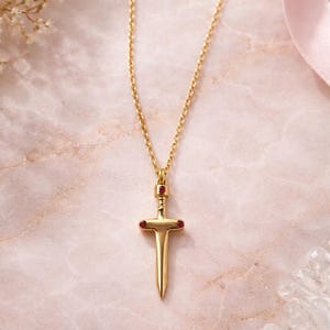Könnte beinhalten: Eine goldfarbene Halskette mit einem Schwertanhänger mit roten Edelsteinakzenten. Der Schwertanhänger hängt an einer zarten Kette. Der Hintergrund ist eine rosa Marmoroberfläche mit floralen und kristallinen Akzenten.