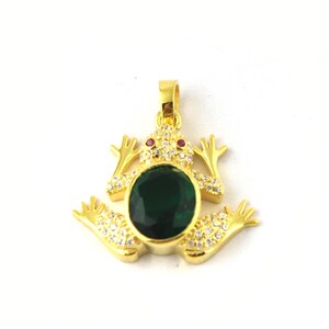 Könnte beinhalten: Goldfarbener Frosch-Anhänger mit einem großen, ovalen, smaragdgrünen Stein in der Mitte. Der Froschkörper ist mit kleinen, klaren Kristallen besetzt und hat rote Akzente für die Augen. Auf weißem Hintergrund.