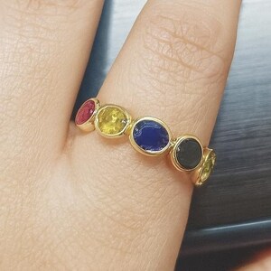 Könnte beinhalten: Ein goldener Ring mit einer Reihe farbiger Edelsteine, darunter Rot, Gelb, Blau und Schwarz. Der Ring befindet sich an einem Finger, im Hintergrund eine Laptop-Tastatur. Die Edelsteine sind oval und in einem goldenen Band gefasst.