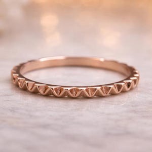 Könnte beinhalten: Roségoldener Ring mit einem sich wiederholenden dreieckigen Muster um das Band. Der Ring ist vor einem weichen, verschwommenen Hintergrund mit Bokeh-Lichtern und weißen Blumen platziert, was eine romantische Ästhetik erzeugt.