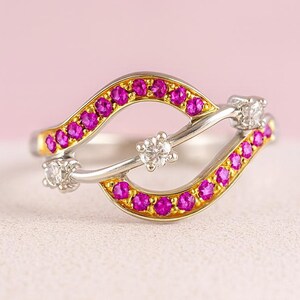 Könnte beinhalten: Ein silberner und goldener Ring mit einem einzigartigen Design. Der Ring hat ein wellenförmiges Muster mit kleinen, leuchtend rosa Edelsteinen und drei klaren, runden Edelsteinen. Der Ring ist vor einem zartrosa Hintergrund platziert.