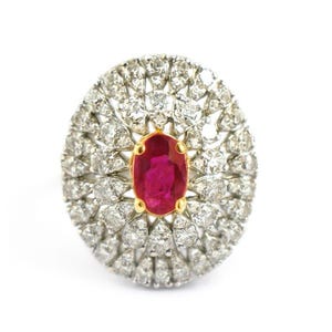 Könnte beinhalten: Ovale Ring mit einem zentralen Rubin-Edelstein, umgeben von einem Kranz aus kleinen, funkelnden Diamanten. Der Ring hat eine Goldeinfassung und ist vor einem weißen Hintergrund platziert.