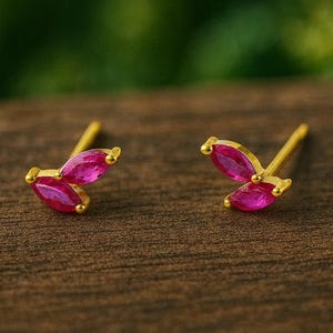 Könnte beinhalten: Ein Paar goldene Ohrstecker mit einem dreiblättrigen Design. Jedes Blütenblatt ist ein lebendiger, marquiseförmiger, fuchsiafarbener Edelstein. Die Ohrringe sind auf einer dunkelbraunen Holzoberfläche mit verschwommenem grünem Hintergrund platziert.