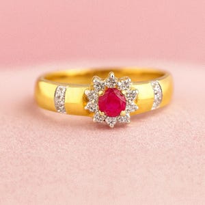 Könnte beinhalten: Ein goldener Ring mit einem zentralen roten Edelstein, der von kleinen weißen Edelsteinen umgeben ist. Das Ringband hat auf jeder Seite des zentralen Steins weiße Edelsteine. Der Ring ist vor einem weichen rosa Hintergrund platziert.