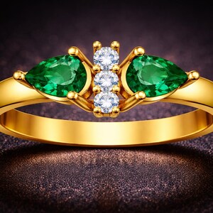 Könnte beinhalten: Ein goldener Ring mit zwei tropfenförmigen Smaragden und drei runden Diamanten. Der Ring hat ein poliertes Goldband und ist vor einem dunklen, strukturierten Hintergrund platziert. Die Edelsteine sind leuchtend grün und klar weiß.