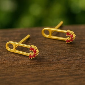 May include: A pair of gold-colored safety pin earrings with red gemstone accents. The earrings are set on a wooden surface with a blurred green background. The earrings are a unique jewelry item.