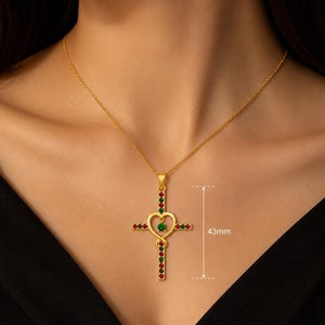 Könnte beinhalten: Eine goldfarbene Kreuz-Halskette mit abwechselnd roten und grünen Steinen an den vertikalen und horizontalen Balken. Eine herzförmige Mitte umschließt einen einzelnen grünen Stein. Eine Grafik zeigt eine Kreuzhöhe von 43 mm an.