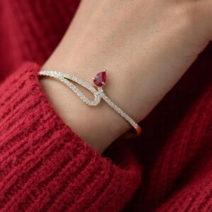 Könnte beinhalten: Ein goldfarbenes Armband mit geschwungenem Design, verziert mit kleinen, funkelnden Kristallen. Ein tropfenförmiger, roter Edelstein ist oben eingesetzt. Das Armband wird an einem Handgelenk vor rotem Strickstoff gezeigt.