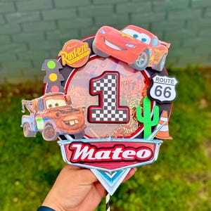 May include: A colorful Cars-themed cake topper featuring Lightning McQueen, Mater, and a Route 66 sign. The number "1" is prominently displayed, along with the name "Mateo". Includes a traffic light and a cactus.