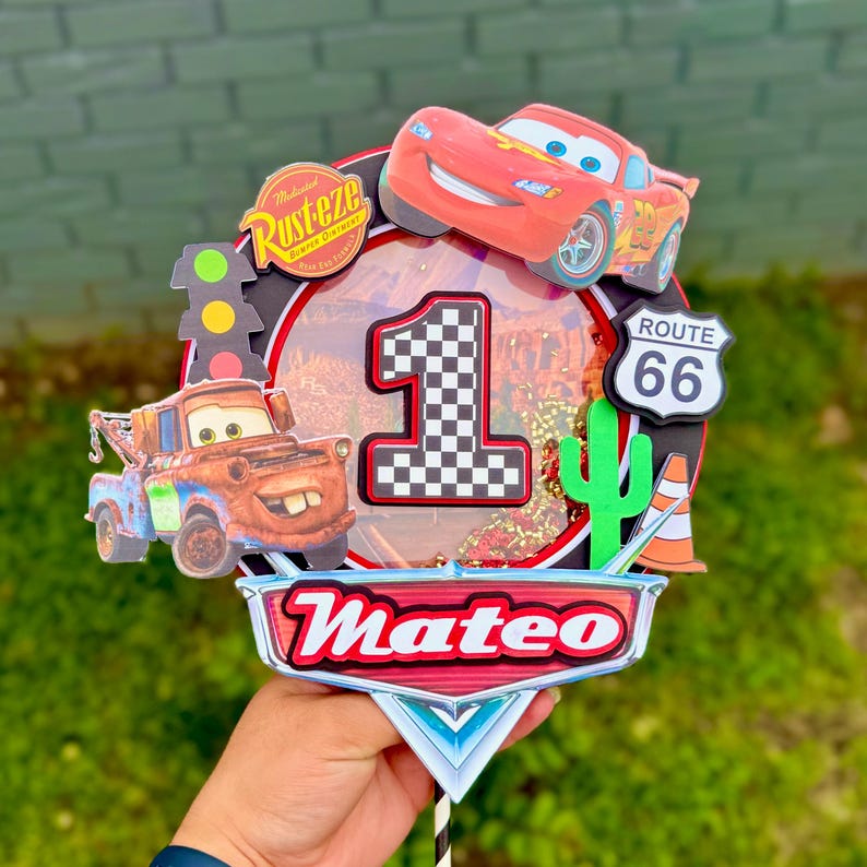 May include: A colorful Cars-themed cake topper featuring Lightning McQueen, Mater, and a Route 66 sign. The number "1" is prominently displayed, along with the name "Mateo". Includes a traffic light and a cactus.