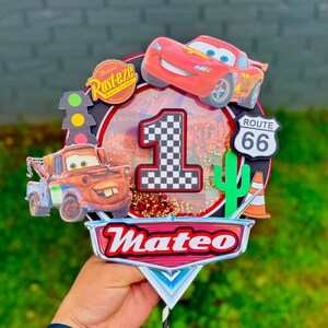 May include: A colorful Cars-themed cake topper featuring Lightning McQueen, Mater, and a checkered number one. Includes a Route 66 sign, a cactus, and the name "Mateo". The topper is designed for a first birthday celebration.