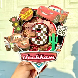 May include: A colorful Cars-themed cake topper featuring Lightning McQueen, Mater, and a checkered number 3. Includes a Route 66 sign, cactus, and the name "Beckham". The topper has a black and white striped stick.