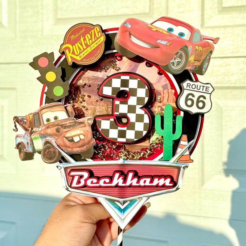 May include: A colorful Cars-themed cake topper featuring Lightning McQueen, Mater, and a checkered number 3. Includes a Route 66 sign, cactus, and the name "Beckham". The topper has a black and white striped stick.
