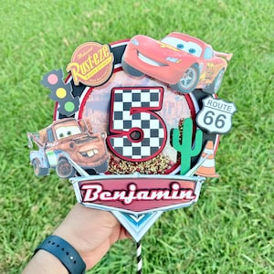 May include: A colorful birthday cake topper featuring Lightning McQueen, Mater, and a Route 66 sign. The number 5 is prominently displayed with a checkered pattern. The name "Benjamin" is also included.
