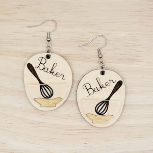 May include: A pair of wooden earrings with a baking theme. Each earring is a light-colored oval with the word "Baker" in a script font. Below the word is a graphic of a whisk inside a bowl. The earrings have silver-colored hooks.