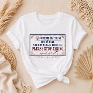 Puede incluir: Una camiseta blanca de manga corta con un gráfico rectangular de borde azul y rojo. El texto dice: OFFICIAL STATEMENT, SHE IS FINE. SHE HAS ALWAYS BEEN FINE. PLEASE STOP ASKING. signed, her. junto a una ilustración de una pluma.