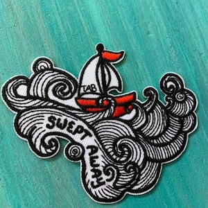 May include: Embroidered patch featuring a sailboat with a red hull and sail, sailing through stylized black and white waves. The patch has the text "SWEPT AWAY" in a decorative font, with the word "TAB" on the sail.