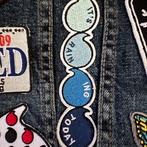 May include: A blue and white embroidered patch with the text "It's Raining Today" on a denim jacket.