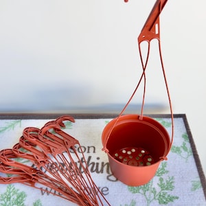 May include: A set of terracotta-colored plastic hanging planters, each featuring a round pot with drainage holes at the base and a long, integrated hook for suspension. One planter is upright, while the others are bundled together in a stack.