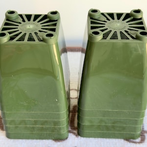 May include: Two stacks of olive green plastic plant pots, each featuring a square base with four raised feet and a radial drainage pattern. The pots have smooth, tapered sides and are nested together in two separate vertical columns.