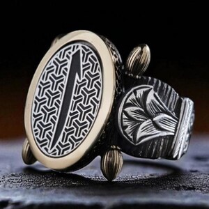 May include: A silver and gold-toned ring with an oval face featuring a geometric pattern and a central vertical design. The ring has decorative elements on the band and small gold-colored accents.