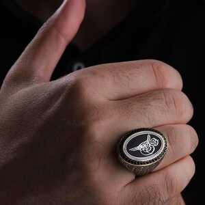 May include: Oval-shaped ring with a black center and a silver design. The ring has a gold-colored band with black accents. The ring is being held up by a hand against a black background.
