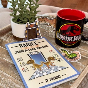 May include: A black Jurassic Park mug with a red interior sits on a wooden tray next to a book and stickers. The mug features the Jurassic Park logo. The book cover has the title "RAIDLE - JURASSIC PARK" and illustrations of dinosaurs.