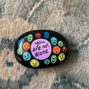 Può includere: Un sasso dipinto di nero con un cerchio di facce sorridenti colorate che circondano un cerchio viola con il testo "you are not alone".