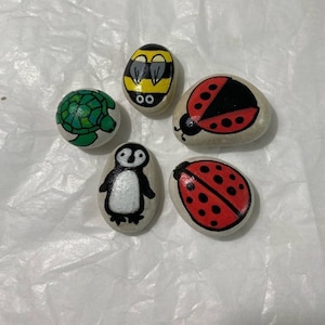 May include: Five painted rocks with different designs. The rocks are painted with a bee, a turtle, a penguin, and two ladybugs.
