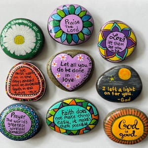 May include: Nine hand-painted rocks with inspirational quotes and floral designs. The rocks feature phrases like "Praise The Lord," "Let all you do be done in love," and "God is good." Colors include green, purple, yellow, and orange.
