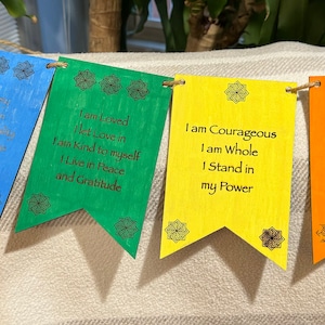 Puede incluir: Una pancarta de colores arcoíris con afirmaciones positivas escritas en cada bandera. Las banderas dicen: "Soy amado, dejo que el amor entre, soy amable conmigo mismo, vivo en paz y gratitud", "Soy valiente, soy completo, me mantengo en mi poder", "Siento mis emociones y mi dolor, reavivo mi pasión, me entrego al momento", "Estoy seguro, confío más, temo menos, estoy centrado y conectado a tierra".