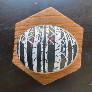 May include: A painted rock with a black and white design of a birch tree forest with two red cardinals perched on branches.