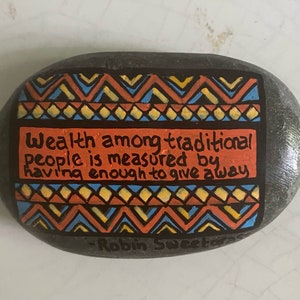 Puede incluir: Una piedra pintada con un patrón geométrico colorido y la cita "La riqueza entre las personas tradicionales se mide por tener lo suficiente para dar." La piedra está firmada "Robin Sweetser".