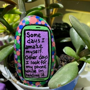 May include: A painted rock with a green, purple, and black phone design. The phone has a white screen with the text "Some days I amaze myself. Other days I look for my phone while I'm holding it."
