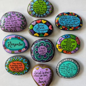Può includere: Una collezione di sassi dipinti con citazioni ispiratrici sul dolore e l'amore. Ogni sasso è decorato in modo unico con disegni floreali colorati e testo scritto a mano. Le citazioni includono frasi come "Il dolore è amore senza un posto dove andare" e "Ricorda".