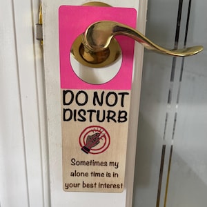 Puede incluir: Un colgador de puerta rosa y de madera con las palabras "DO NOT DISTURB" en negro. El colgador tiene un gráfico de una mano sosteniendo un lápiz roto y el texto "Sometimes my alone time is in your best interest."