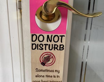 Puede incluir: Un colgador de puerta rosa y de madera con las palabras "DO NOT DISTURB" en negro. El colgador tiene un gráfico de una mano sosteniendo un lápiz roto y el texto "Sometimes my alone time is in your best interest."