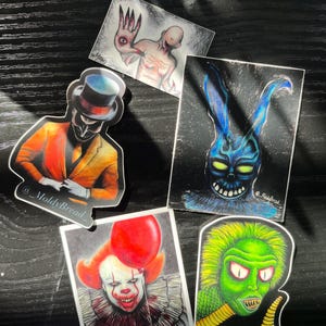 May include: A collection of stickers featuring horror-themed illustrations. The stickers include a clown with a red balloon, a blue rabbit with glowing eyes, a green creature, and other eerie figures. The stickers are on a dark surface.