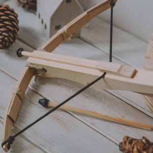 May include: A wooden toy crossbow with a black string and a small arrow. The crossbow is made of light-coloured wood and has a simple design. The background includes a small white house and a pinecone.