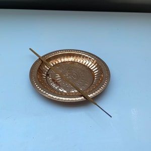May include: A small, round, copper incense burner with a raised design of the Om symbol. A brown incense stick is lying next to the burner.