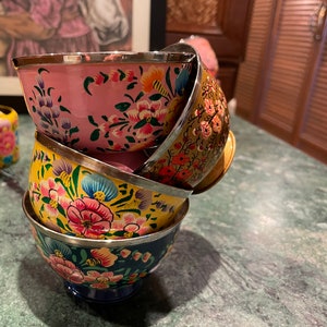 May include: A stack of four hand-painted bowls with floral designs. The bowls are nested inside each other, with the top bowl being pink, the next yellow, then blue, and the bottom bowl is not visible. The bowls have a silver rim.