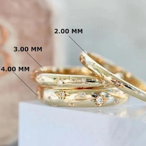 Könnte beinhalten: Goldfarbene Ringe mit Stern- und Edelsteinbesatz. Die Ringe sind gestapelt und zeigen Breiten von 2,00 mm, 3,00 mm und 4,00 mm. Die Ringe haben eine polierte Oberfläche und sind vor einem weichen, neutralen Hintergrund platziert.