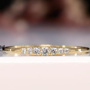 Puede incluir: Un delicado anillo de oro con una fila de pequeñas piedras preciosas claras engastadas en la parte superior. El anillo tiene una banda delgada y un diseño simple y elegante. El anillo se muestra sobre una superficie blanca.