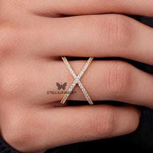 May include: A gold ring in an X-shaped design, set with small, clear gemstones. The ring is on a finger, with the hand and part of a black sleeve visible. The ring is from Stella Jewelry.