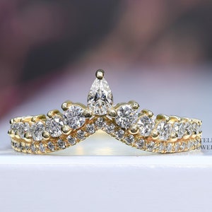 Pode incluir: Um anel de ouro com um diamante em forma de pêra no centro e diamantes menores ao redor. O anel é projetado em forma de coroa.