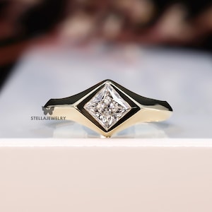 May include: A gold ring with a square-cut diamond center stone. The diamond is set in a geometric bezel setting. The ring has a polished finish and is displayed on a white surface. The ring is a piece of jewelry.