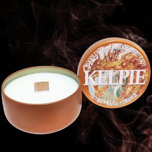 May include: A copper-colored candle tin with the word "KELPIE" in white lettering. The tin is open, revealing a white candle with a wooden wick. The lid displays the words "WOODLUNGS HANDCRAFTED SOY" and the scent notes: thyme, basil, and orange.