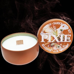 May include: A brown soy candle in a round tin with a wooden wick. The candle has a white wax and a green rim. The lid of the tin is orange with the word "PIXIE" and the words "Eucalyptus, Aloe, Spearmint".
